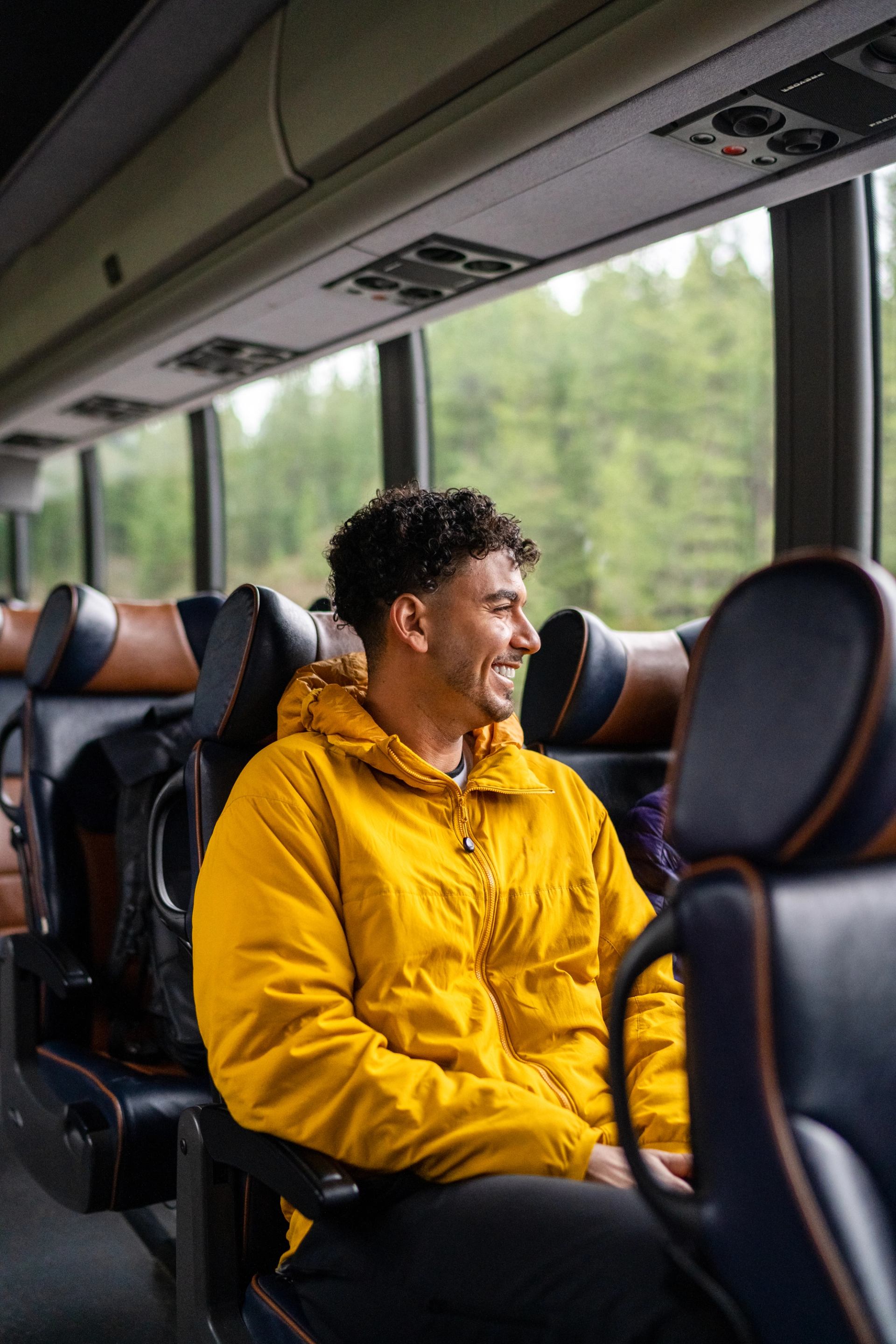 Passenger in a yellow jacket seated on a shuttle bus near a window, with blurred forest scenery passing outside; the interior shows rows of dark leather seats, overhead panels, and natural light coming through large windows.