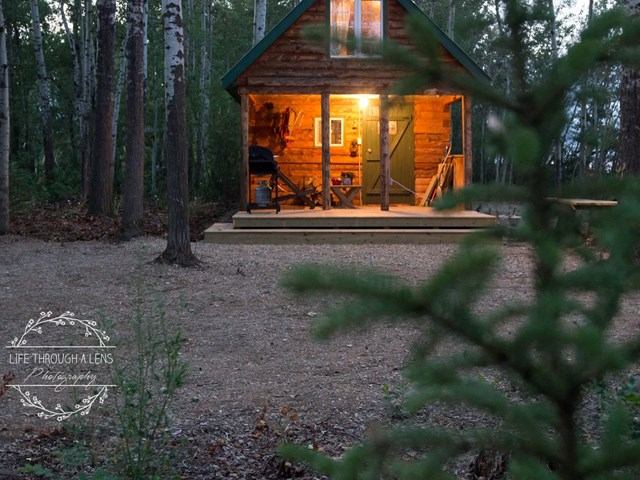 Wood cabin with lit porch, steps, and outdoor seating in a forest clearing at dusk.