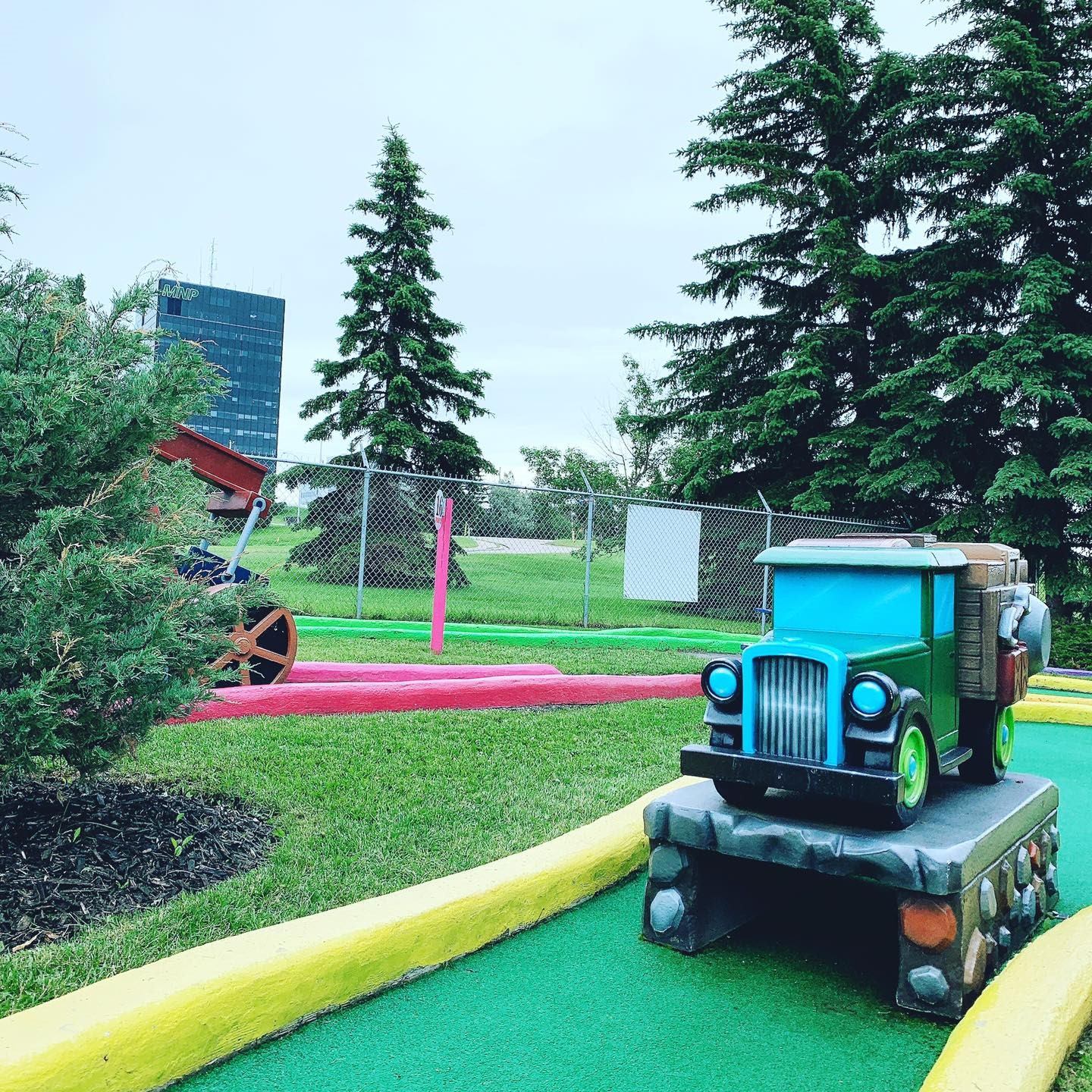 Mini golf course with painted barriers, net fence, trees, and a green toy truck.