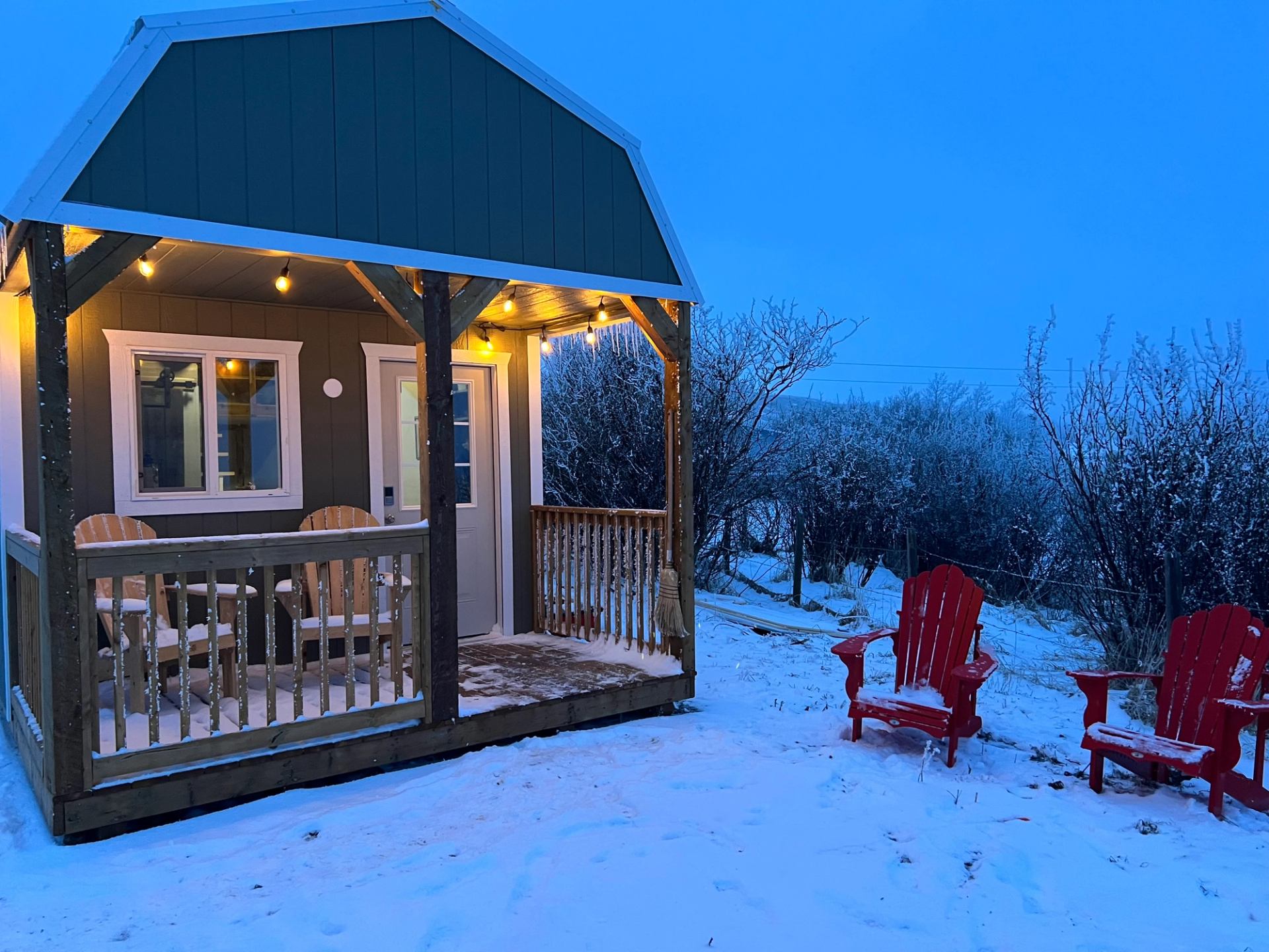 Small cabin with porch lights, snow-covered ground, and two red chairs outside at dusk.