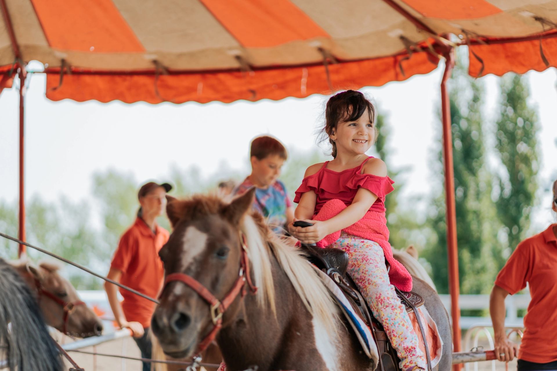 Child riding a pony at Spruce Meadows under a striped tent, with attendants and other riders nearby.