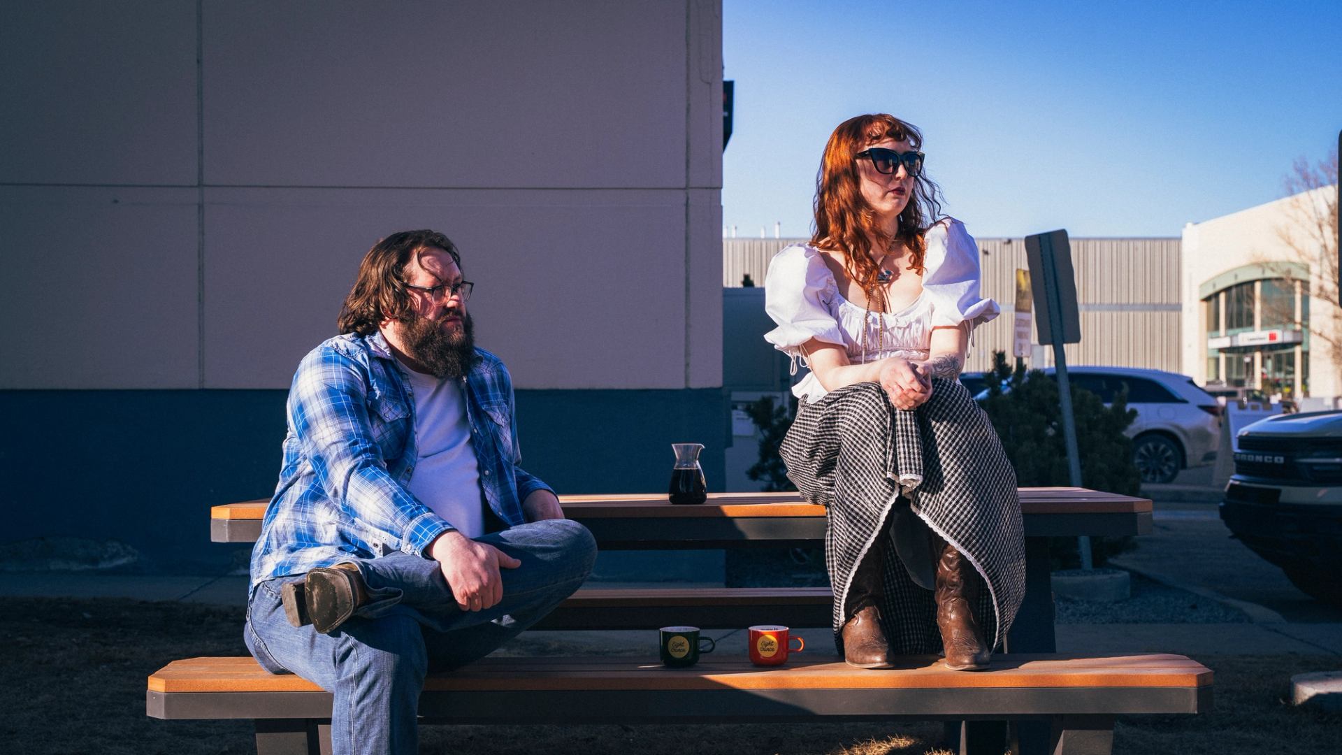 Two people sitting on a picnic table outdoors with coffee mugs and a carafe in a sunny western‑style setting.