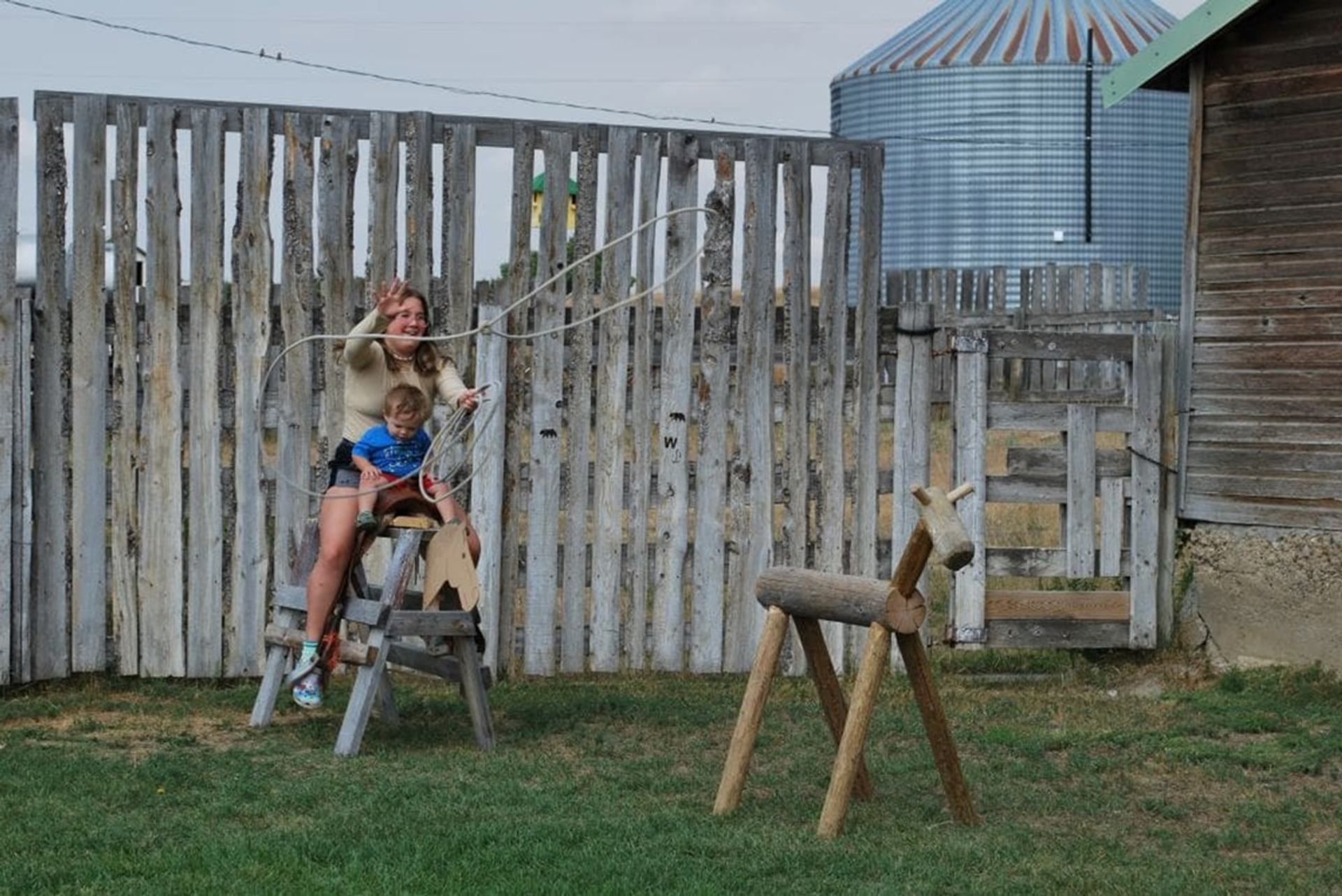 Person and child on wooden horse practise lassoing wooden calf in fenced farmyard.