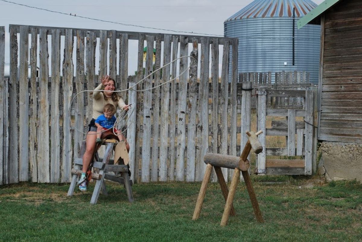 Person and child on wooden horse practise lassoing wooden calf in fenced farmyard.