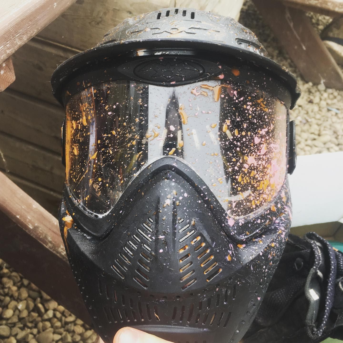 Paintball mask with clear visor splattered with orange and pink paint.