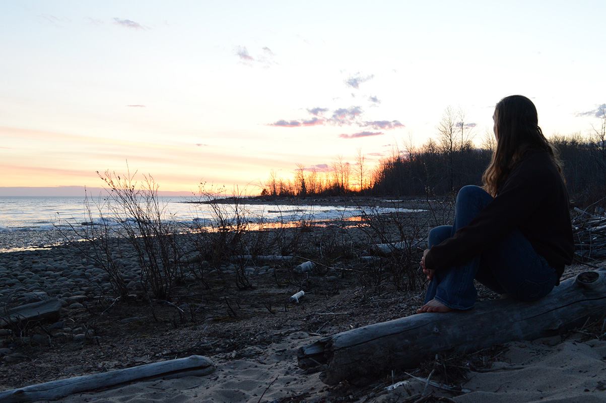 A woman sits on a driftwood log, admiring the sunset.