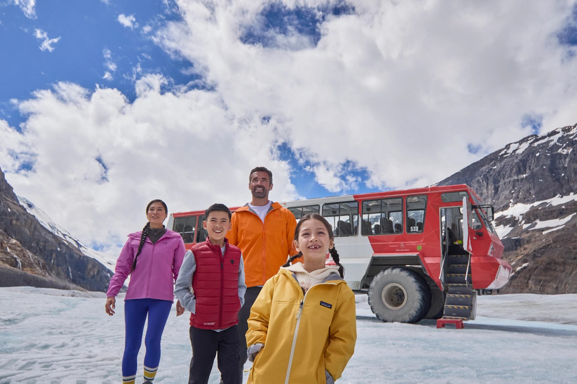 Group of four people on snowy glacier with red tour bus and mountains behind.