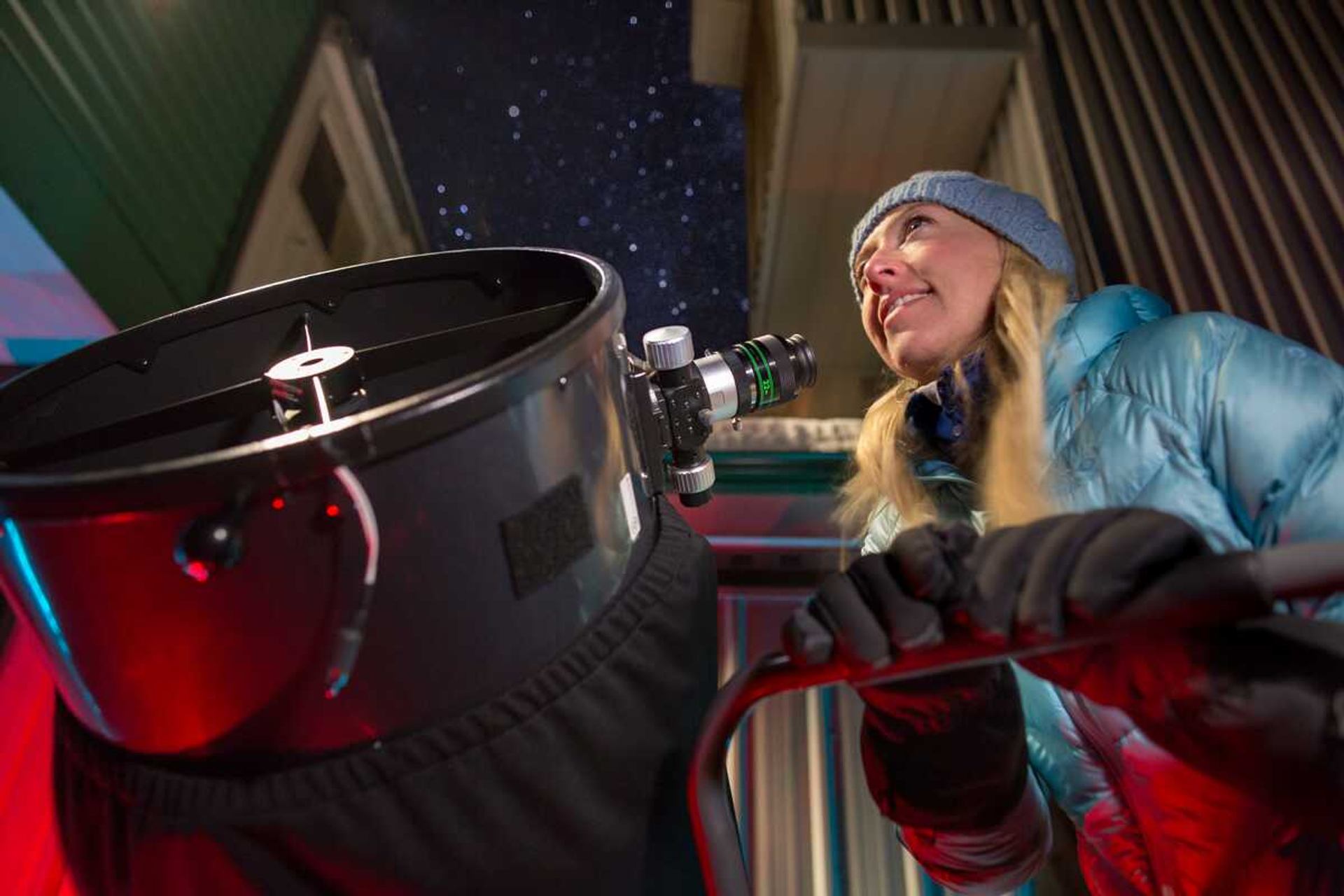 Person in winter clothing adjusts large telescope under starry night sky between buildings.