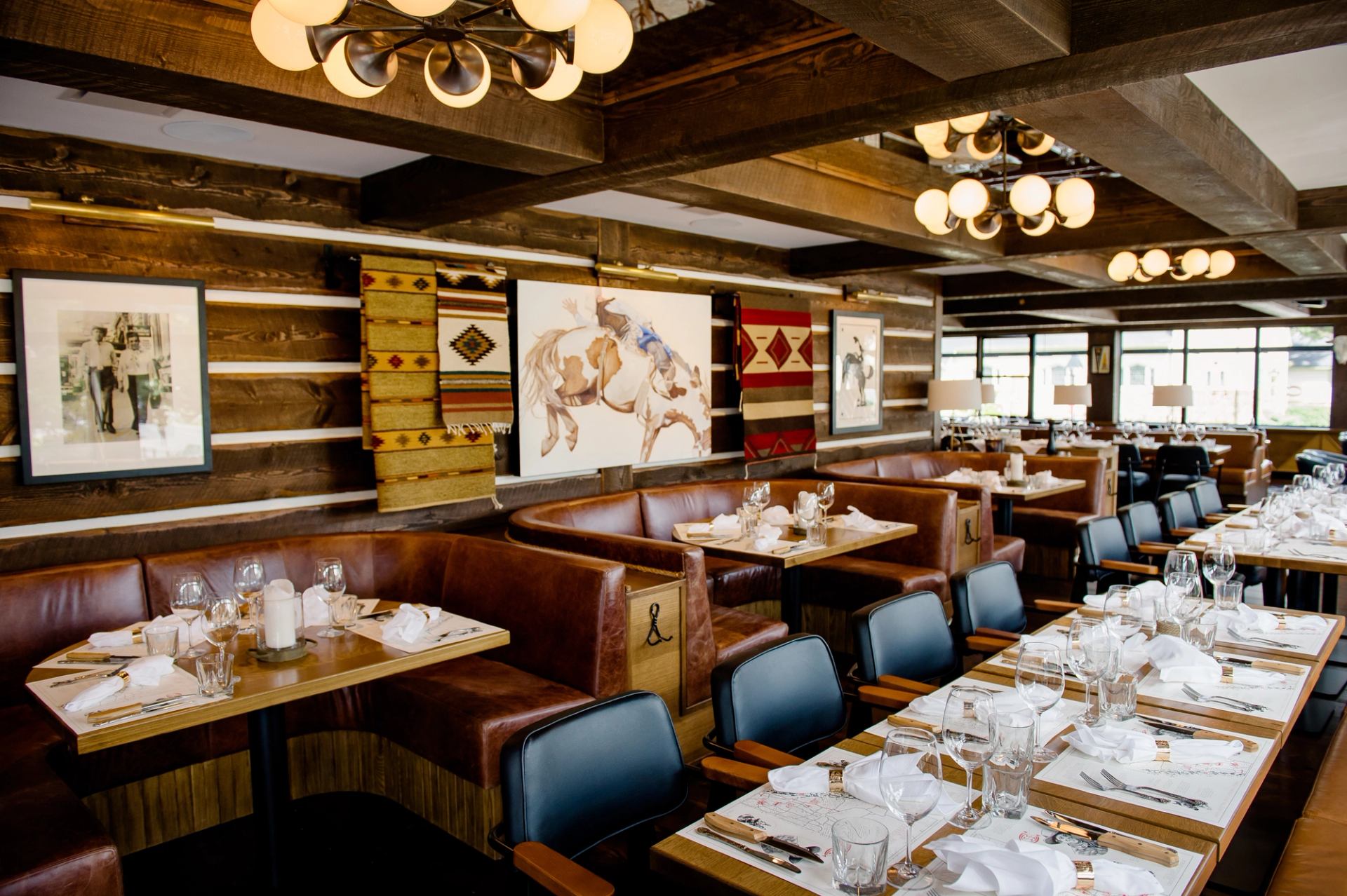 Rustic restaurant interior with wood log walls, exposed beams, leather booths, set tables, and Western-themed art.