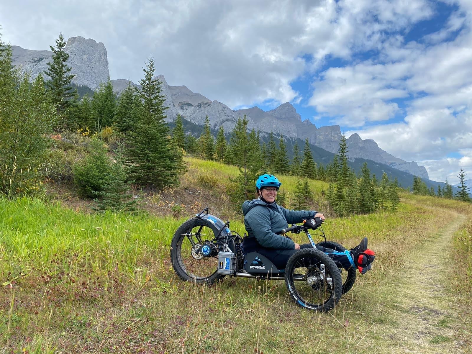 Person riding adaptive trike on grassy trail with mountains and trees.