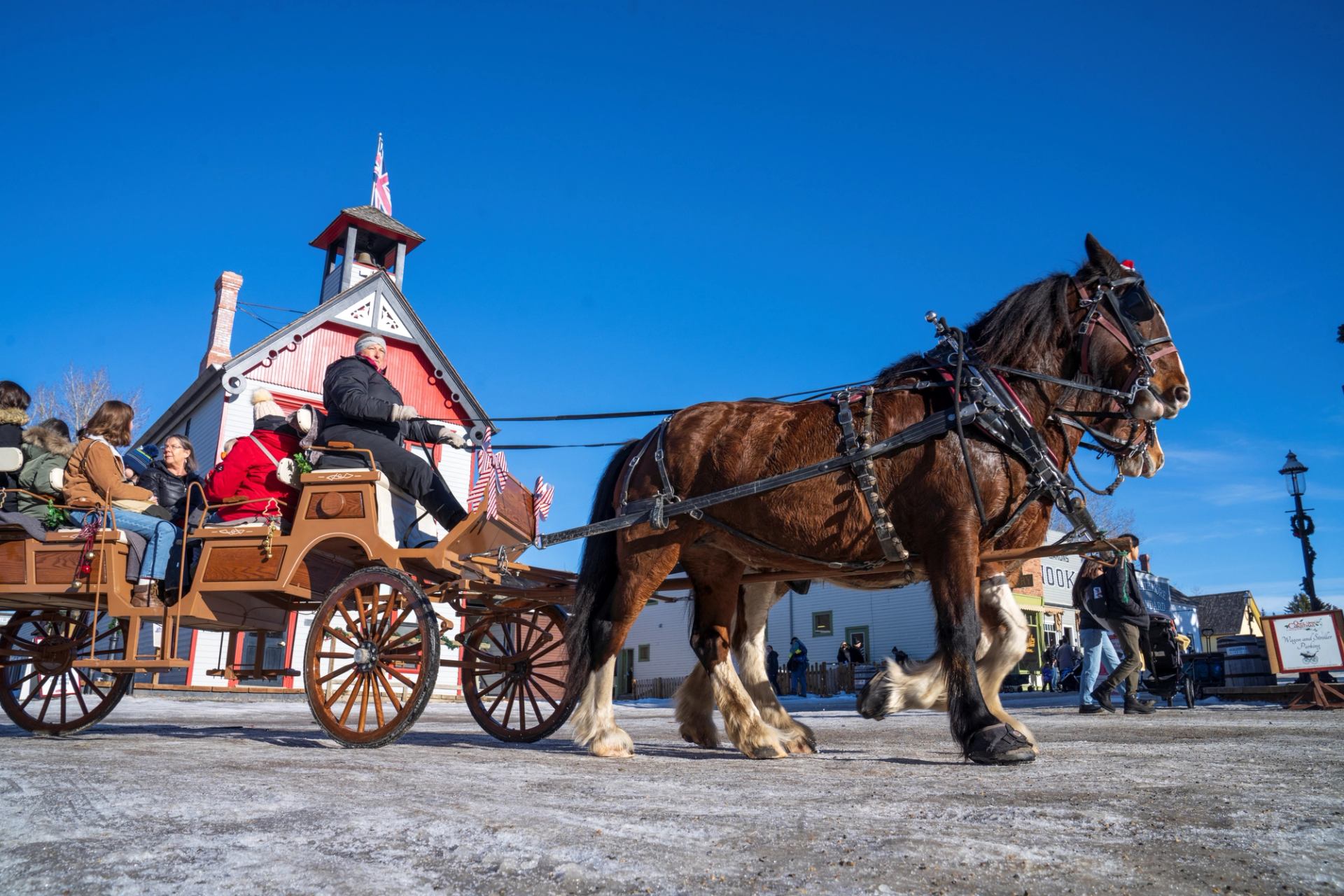 Horse-drawn wagon carries visitors past a red heritage building on a clear winter day.