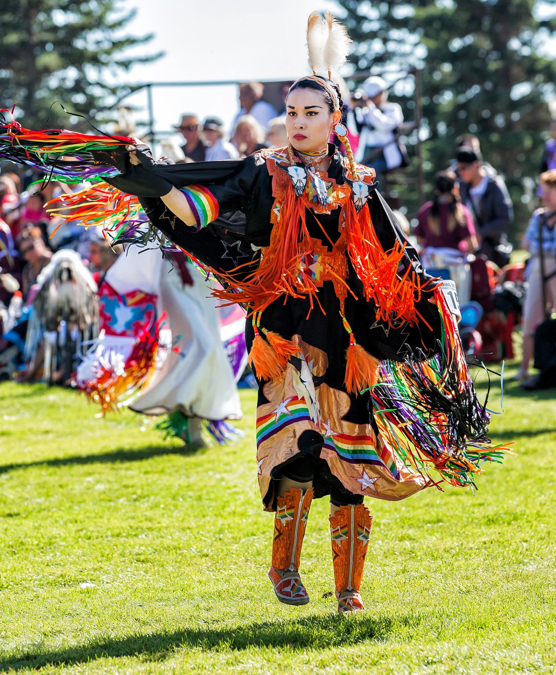 Powwow dancer in colourful regalia performs on grass, with spectators and trees in background.