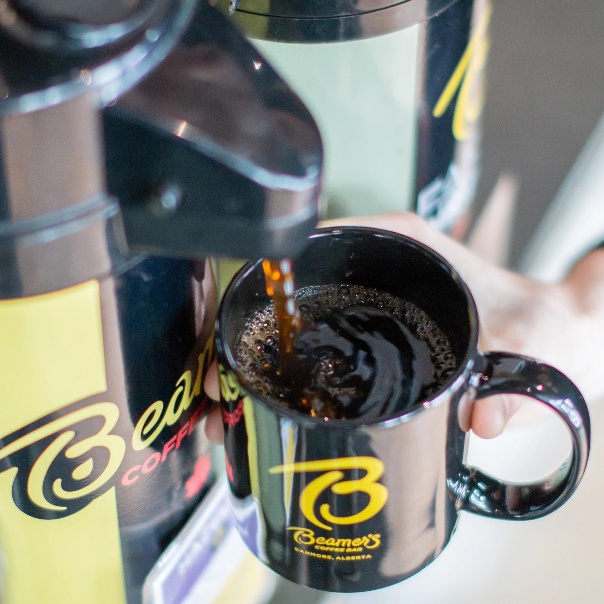 Coffee dispenser pouring dark coffee into a Beamer’s Coffee mug held by hand.
