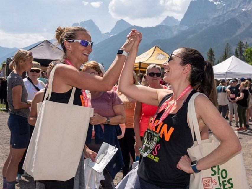 Runners in sleeveless shirts high-five at an outdoor race with mountain peaks in background.