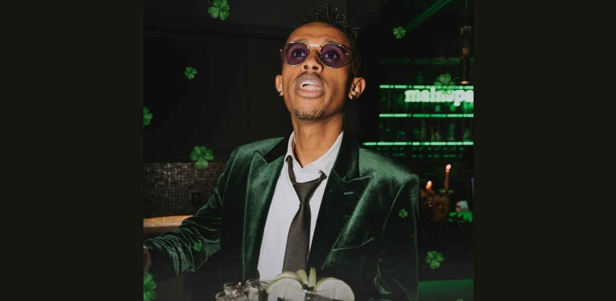 Person in a green velvet jacket stands at a bar with cocktails, surrounded by St. Patrick’s Day décor and shamrocks.