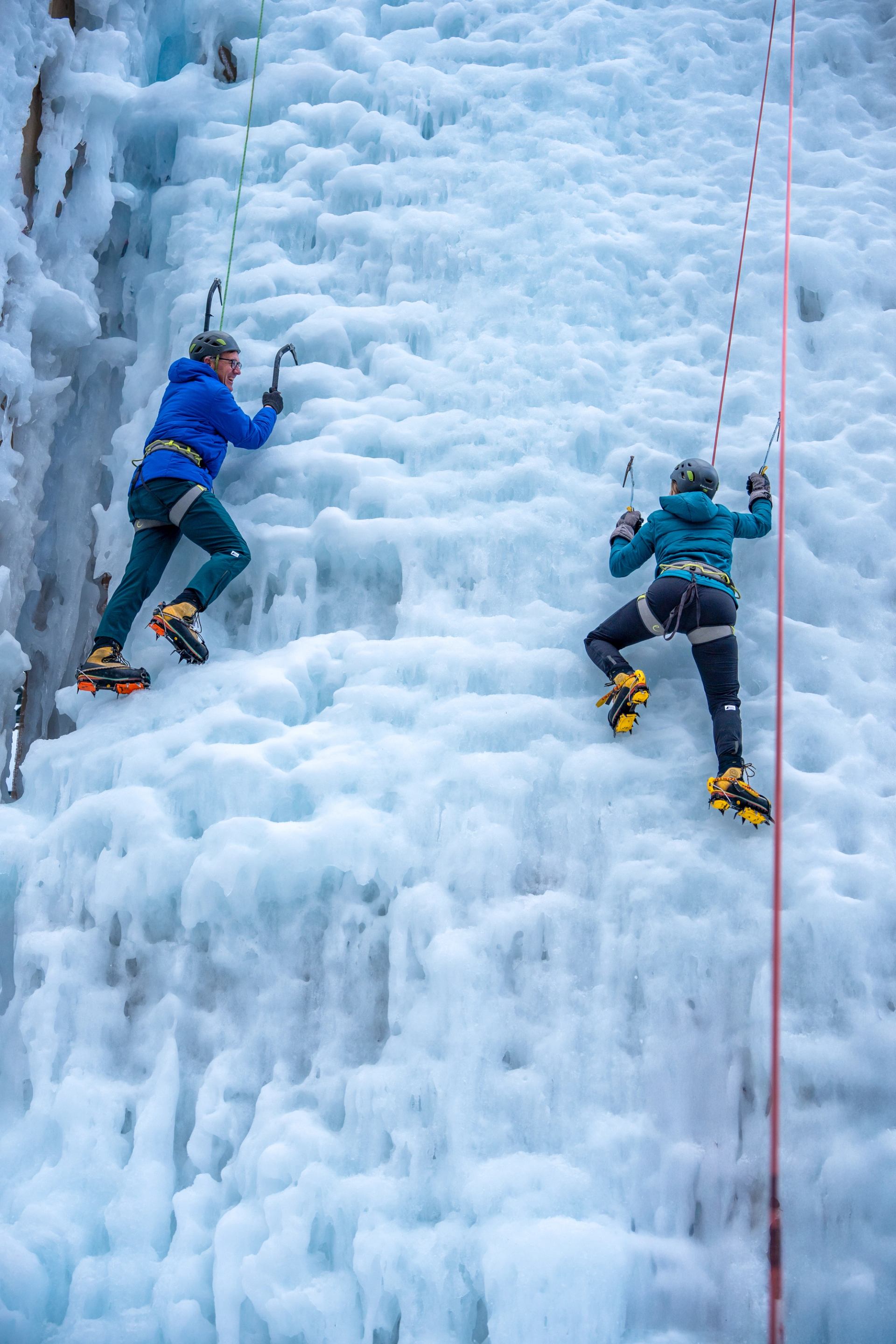 Two climbers scaling icy wall with ropes, axes, and winter gear on frozen waterfall.