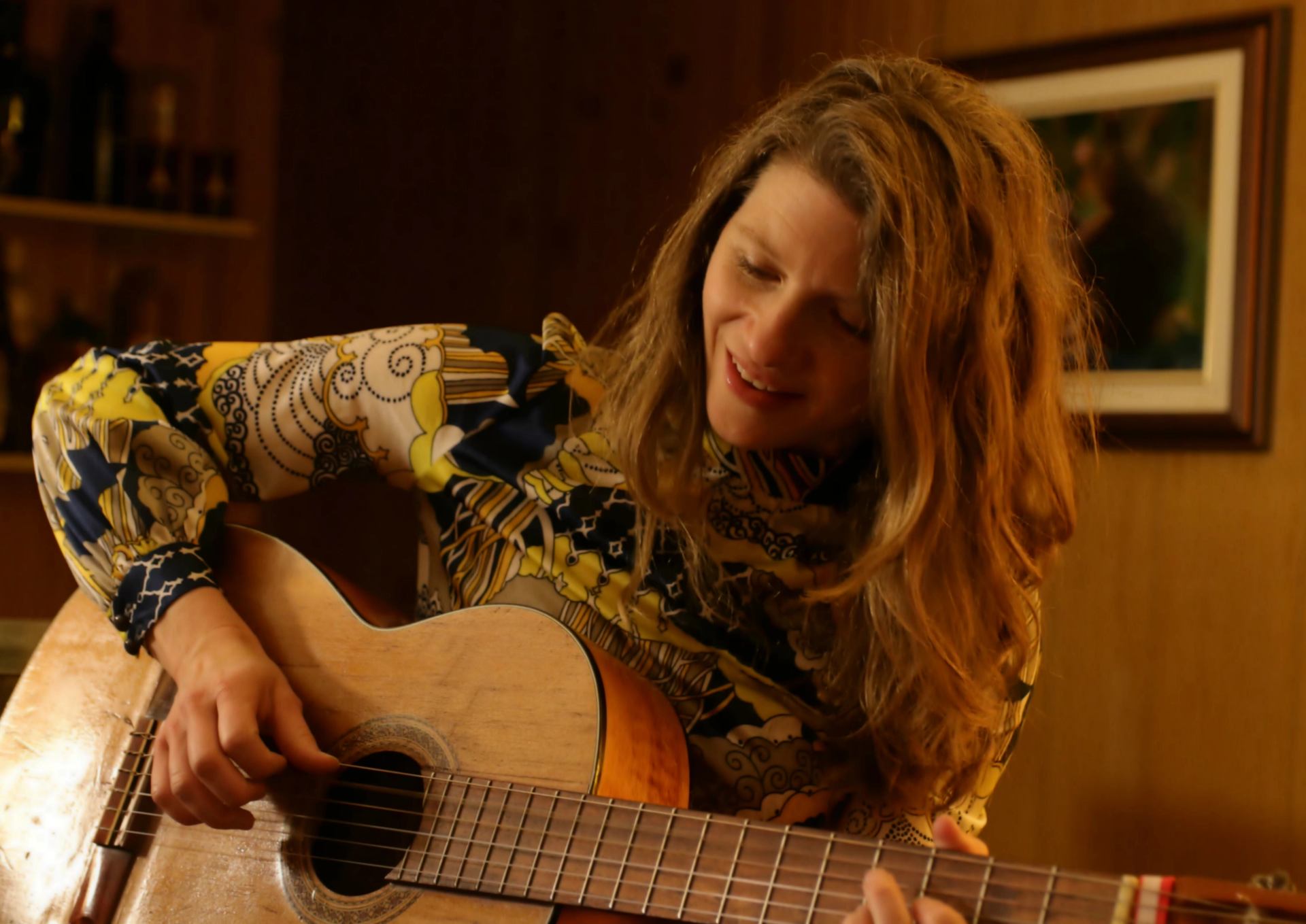 Person with long hair plays an acoustic guitar indoors near a framed picture.