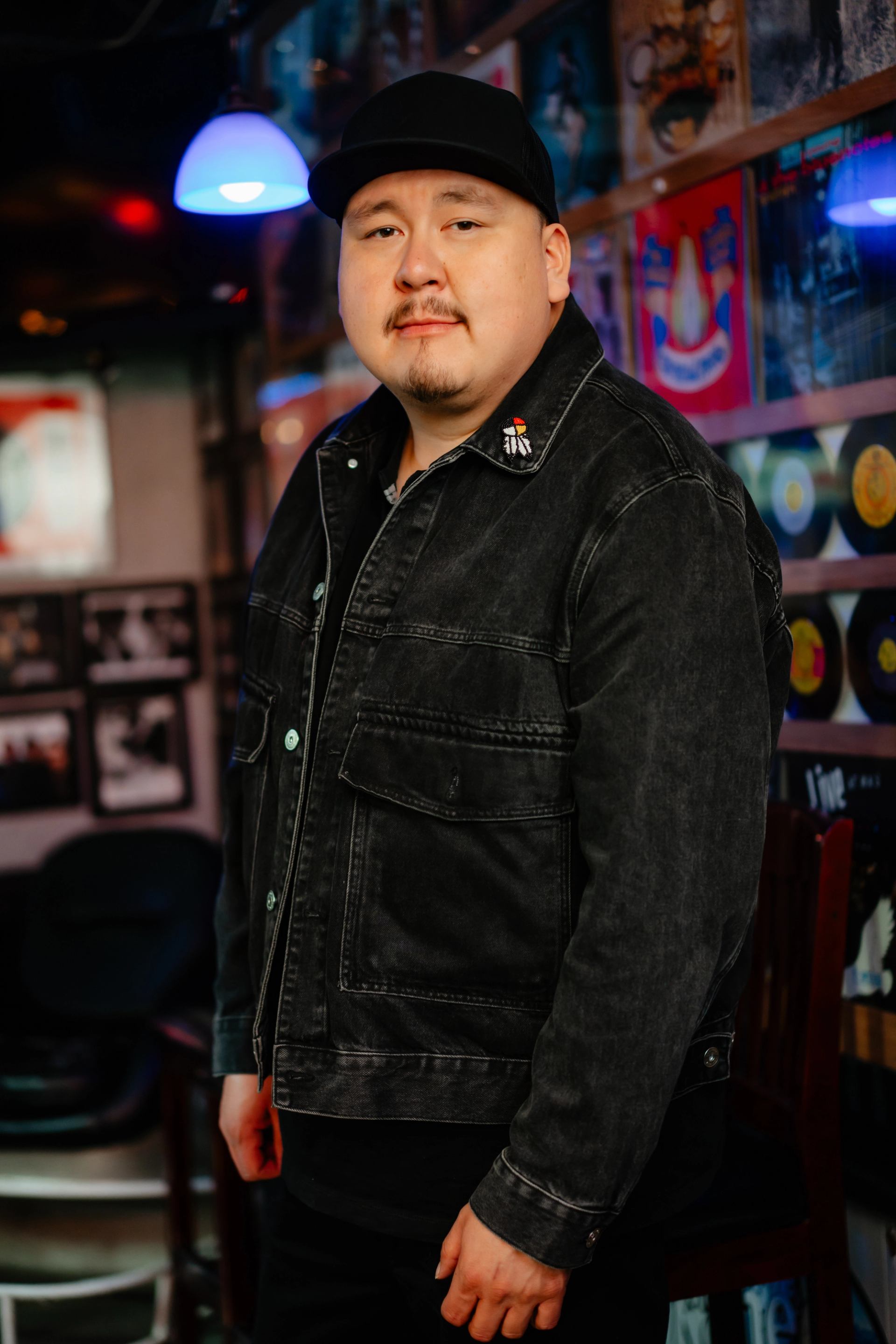 Person in black jacket and cap stands indoors before wall of colourful album covers.