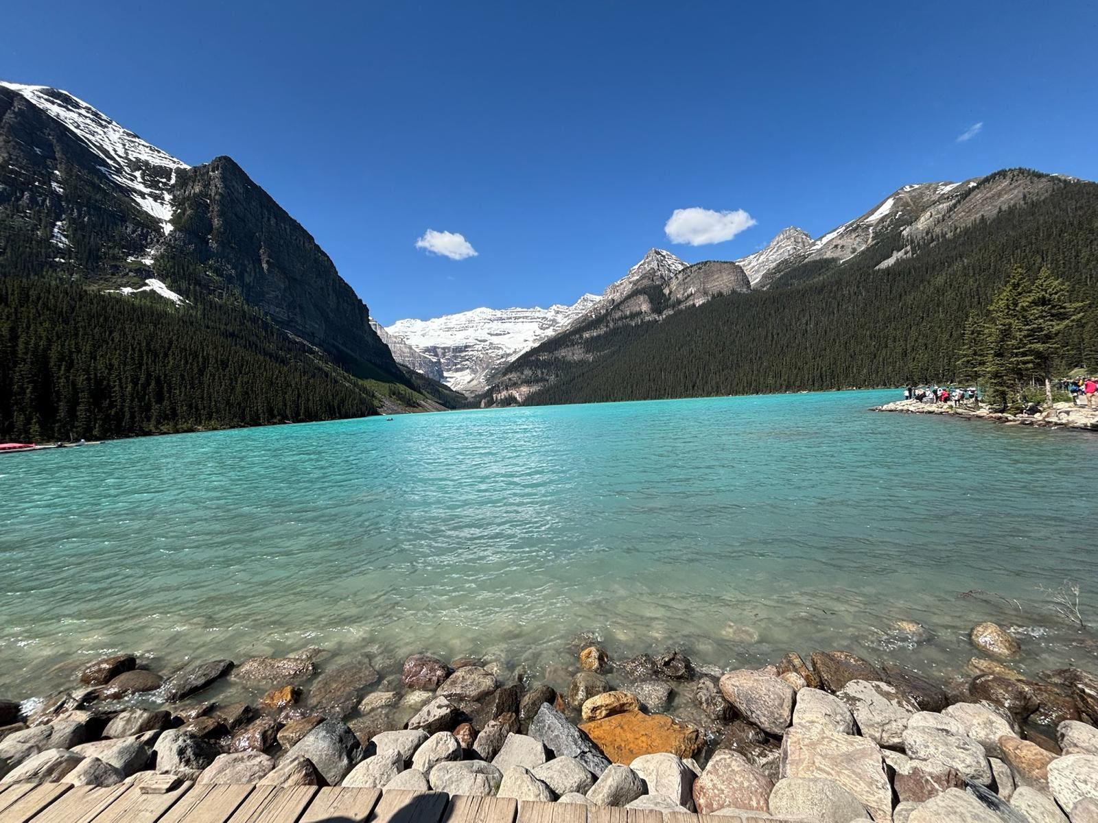 Turquoise lake with rocky shore, forested slopes, and snow-capped mountain peaks.