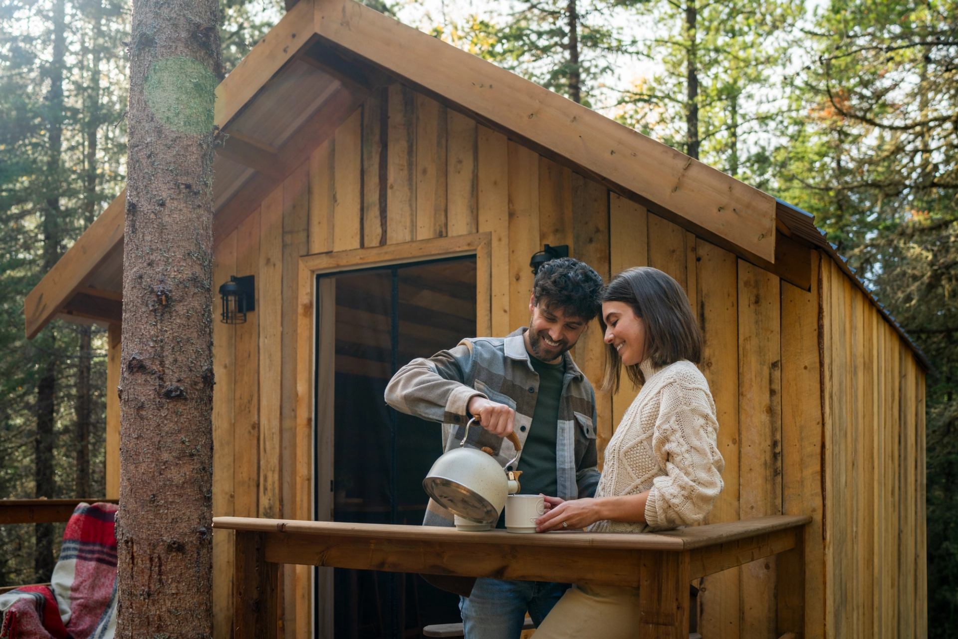 A man pours from a kettle, while on the deck of a small cabin with a woman.
