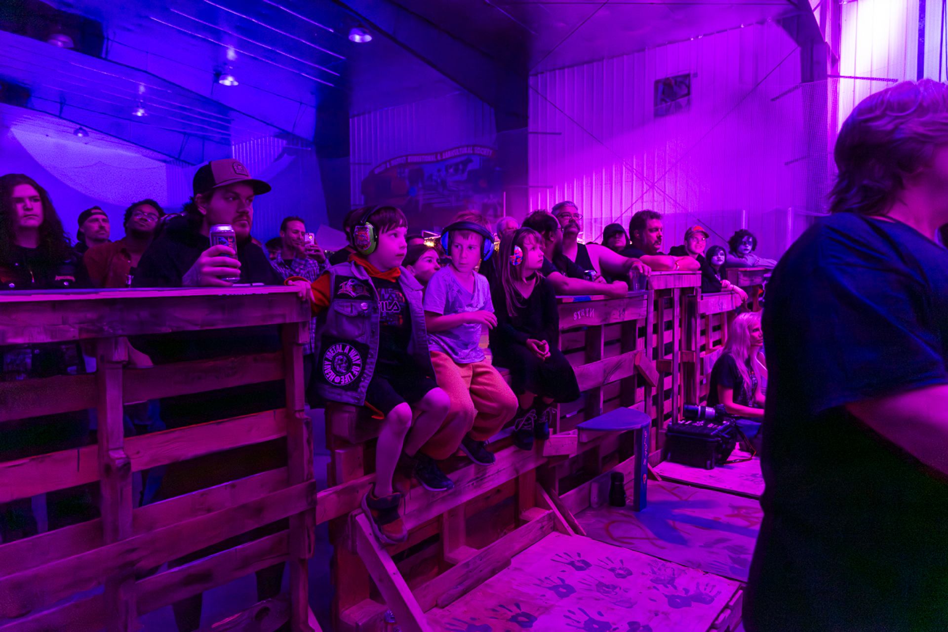 Decimate Metalfest crowd stands behind a barrier under purple lights, with young fans wearing ear protection watching stage.