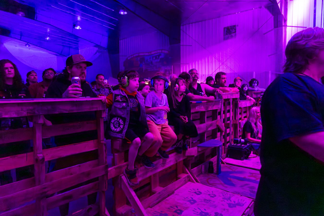 Decimate Metalfest crowd stands behind a barrier under purple lights, with young fans wearing ear protection watching stage.