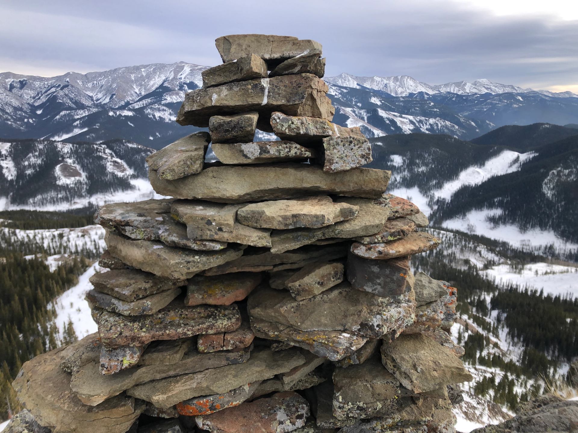 Stacked stone cairn on a snowy mountain ridge with distant peaks under a cloudy winter sky.