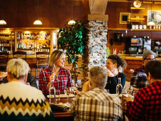 Group of people dining and talking at a cozy restaurant with wood décor and warm lighting.