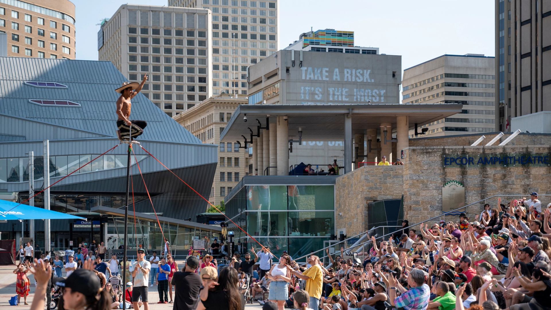 Street performer balances on tall pole as large crowd watches in urban square.