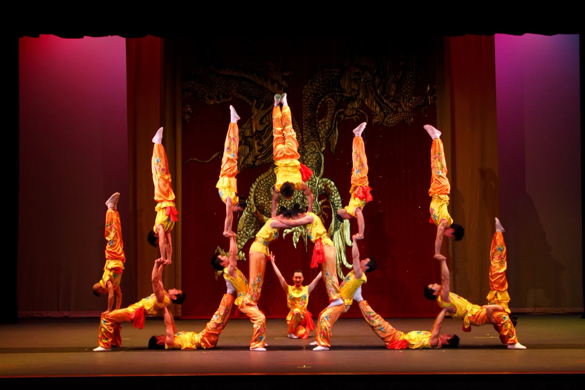 Acrobats in yellow costumes form a balanced human pyramid on a red theatre stage.