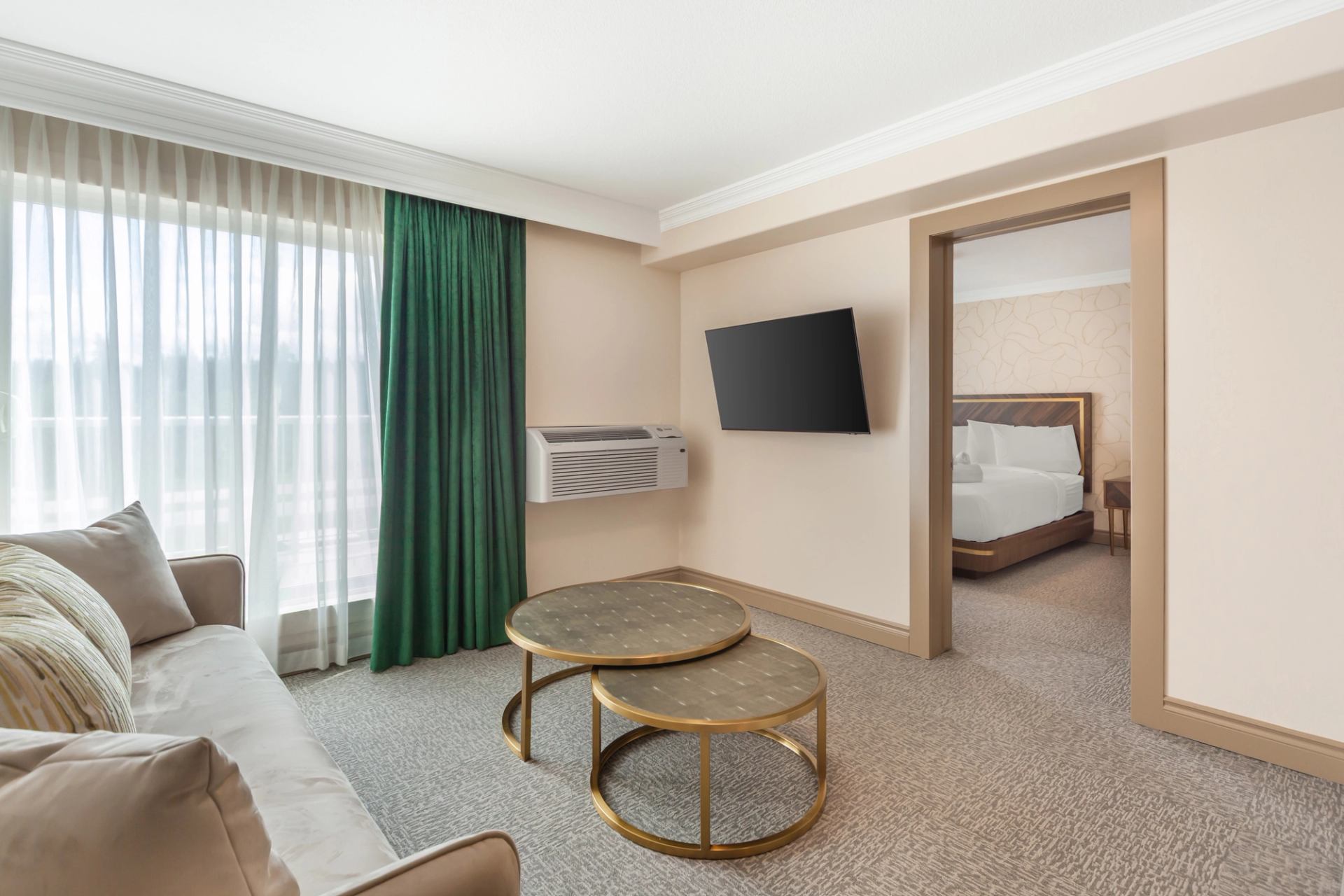 Spacious hotel suite with a sofa, nested coffee tables, wall-mounted TV, and a view into the bedroom.
