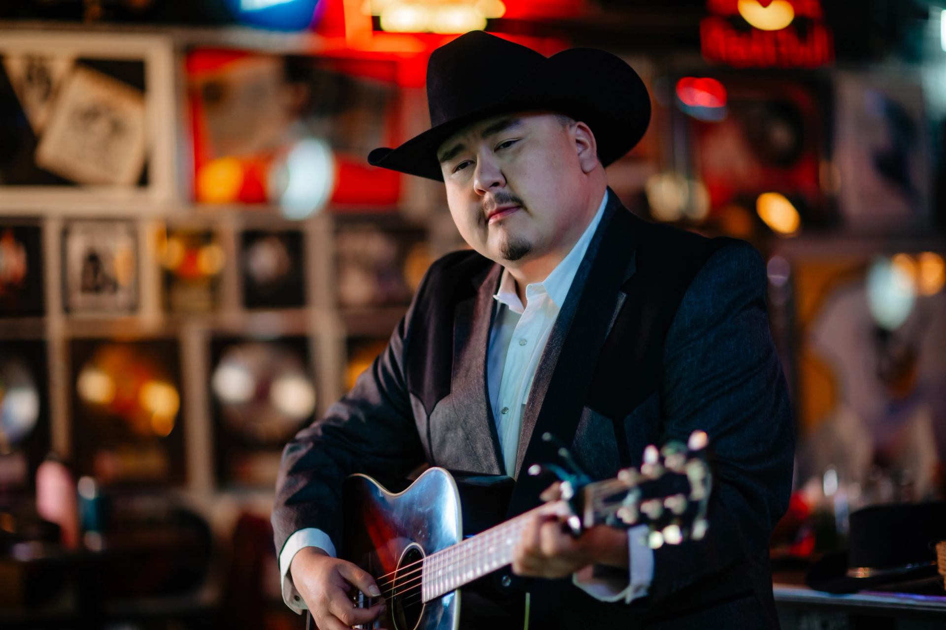 Musician in dark suit and cowboy hat plays acoustic guitar in neon-lit bar.