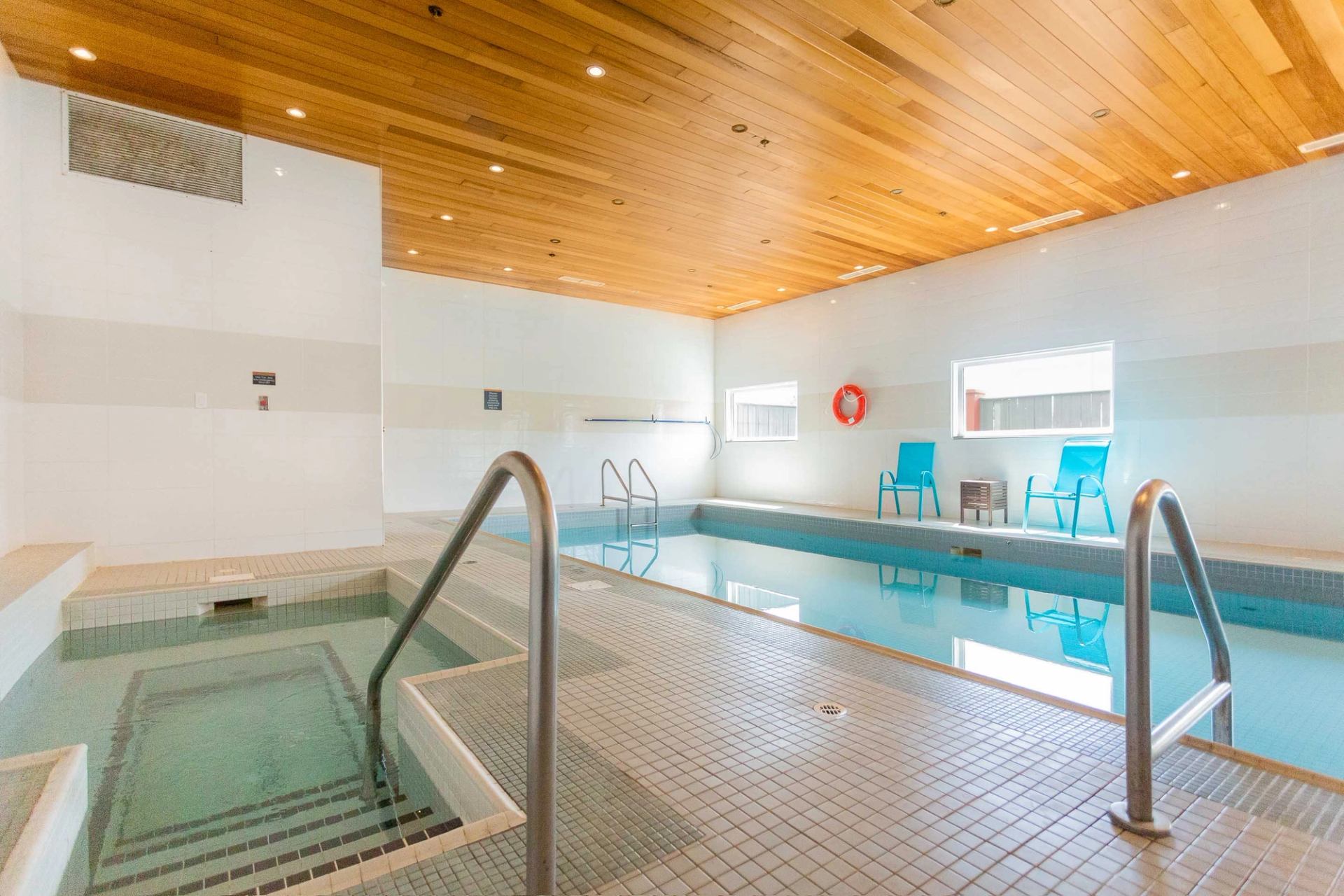 Indoor pool and hot tub area with tiled deck, wooden ceiling and blue chairs.