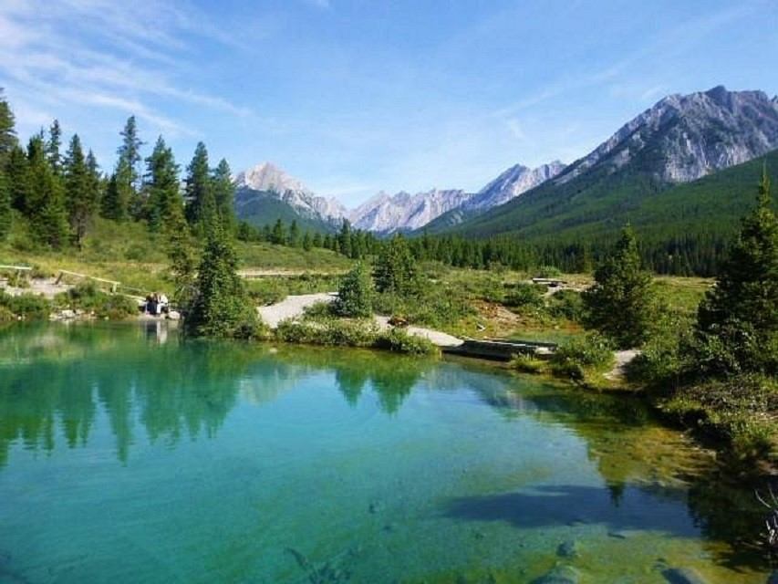 Turquoise pond beside trail, with evergreen forest and rocky mountain peaks.