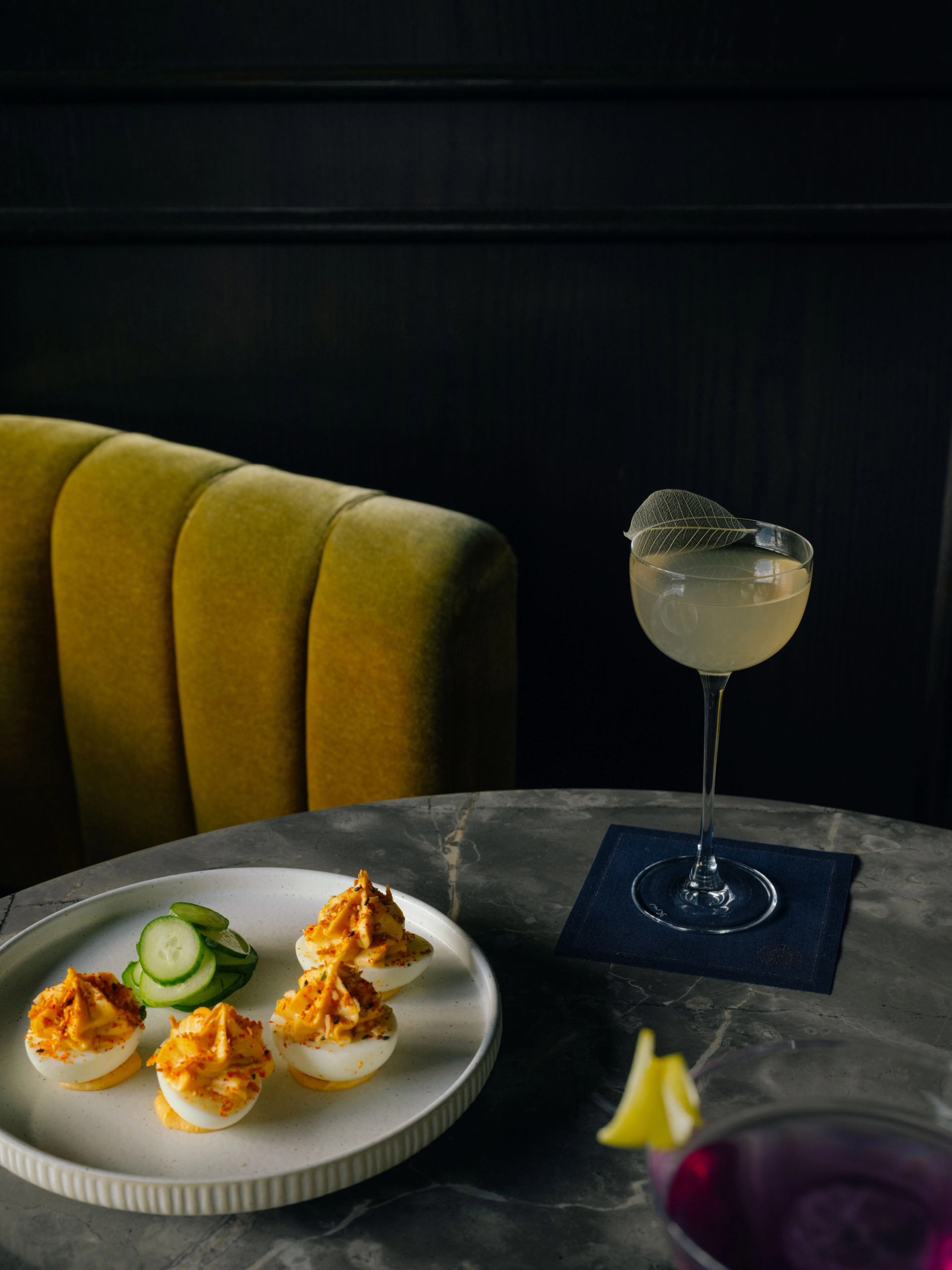 Cocktail and small bites on a marble table with a yellow velvet booth in the background.