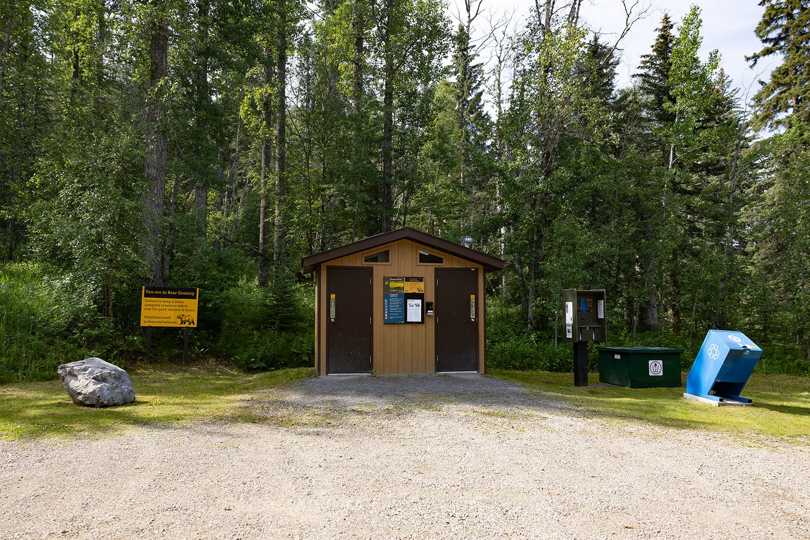 Wood utility building, yellow camping sign, recycle bin and gravel driveway in forest campground.