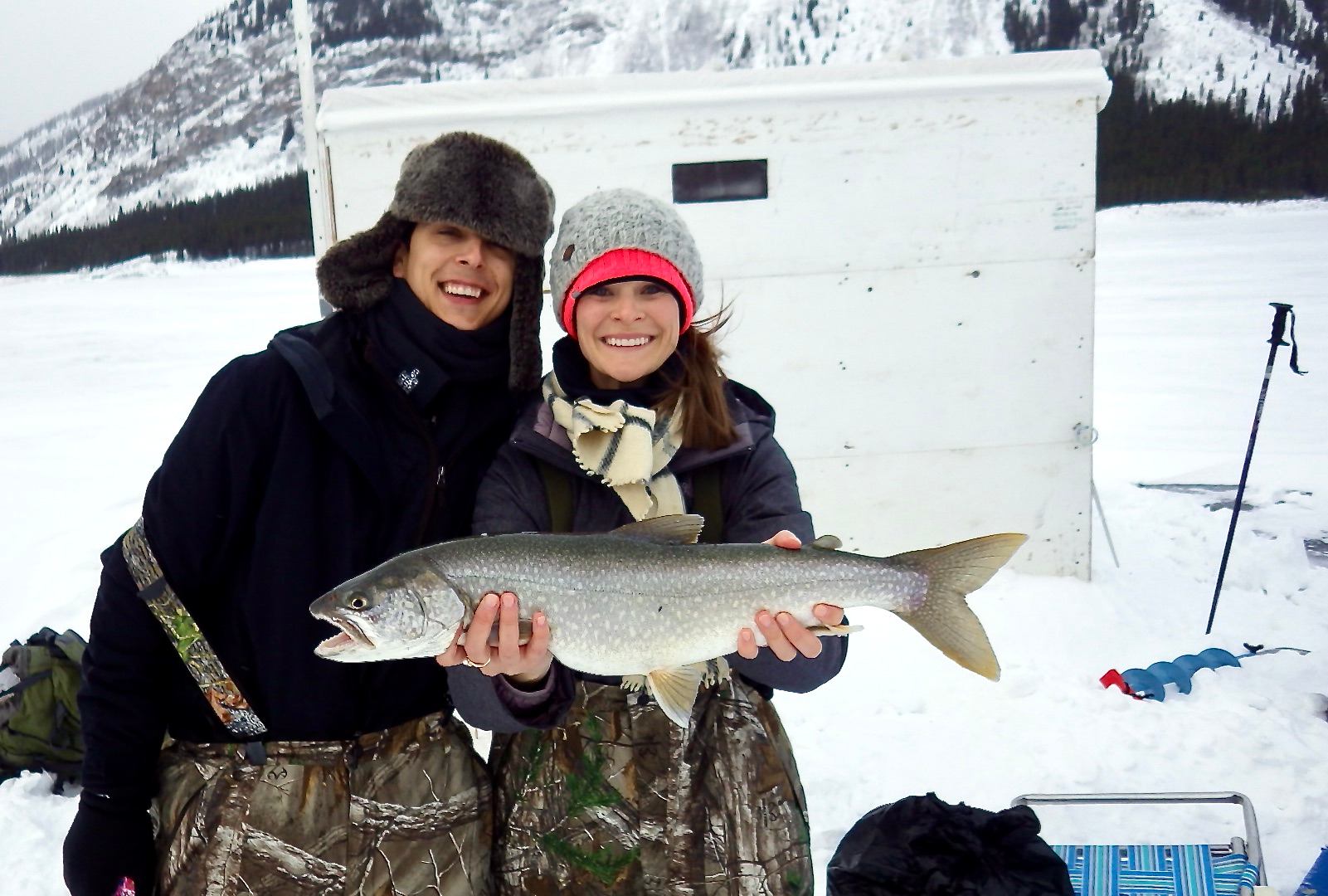 Two bundled people hold a large fish on snowy ice near a white shack.