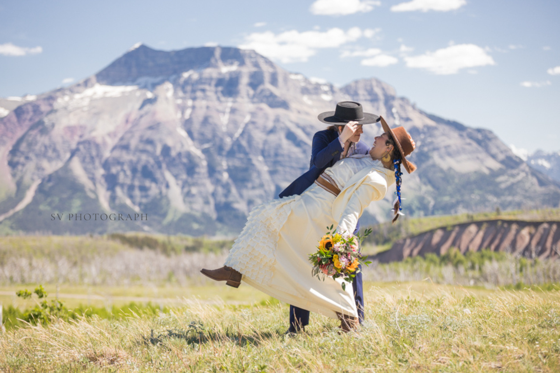 A couple embraces in a grassy field as one is dipped back, holding flowers, with mountains behind.