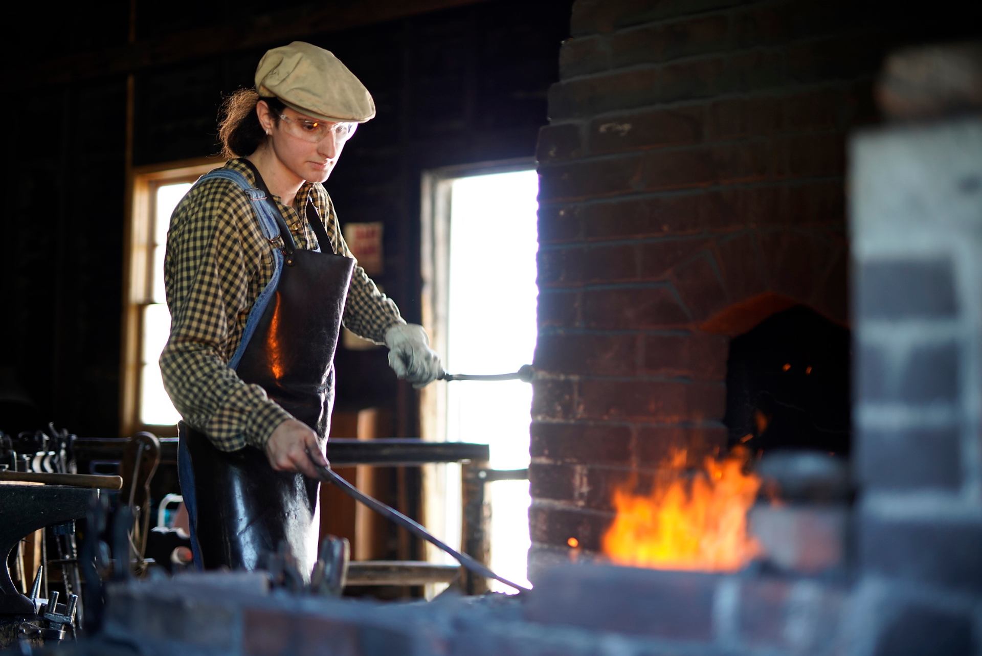 Blacksmith wearing safety gear heats metal rod in forge with bright orange flames.