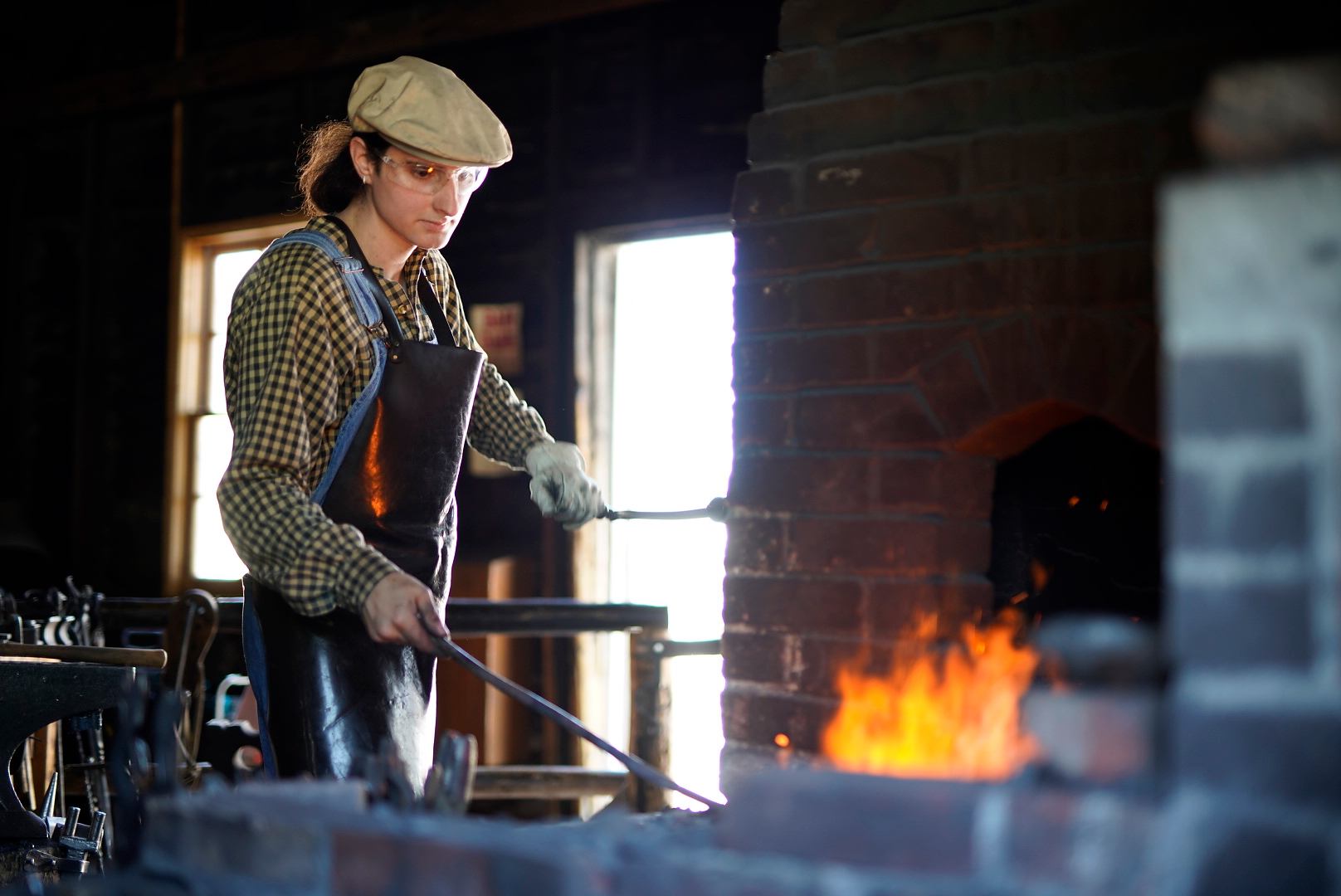 Blacksmith wearing safety gear heats metal rod in forge with bright orange flames.