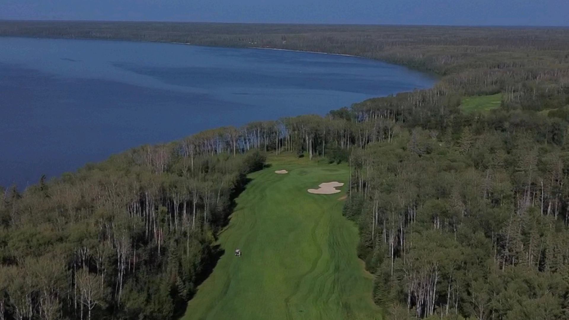 Golf fairway with sand bunkers runs through forest beside a large lake.