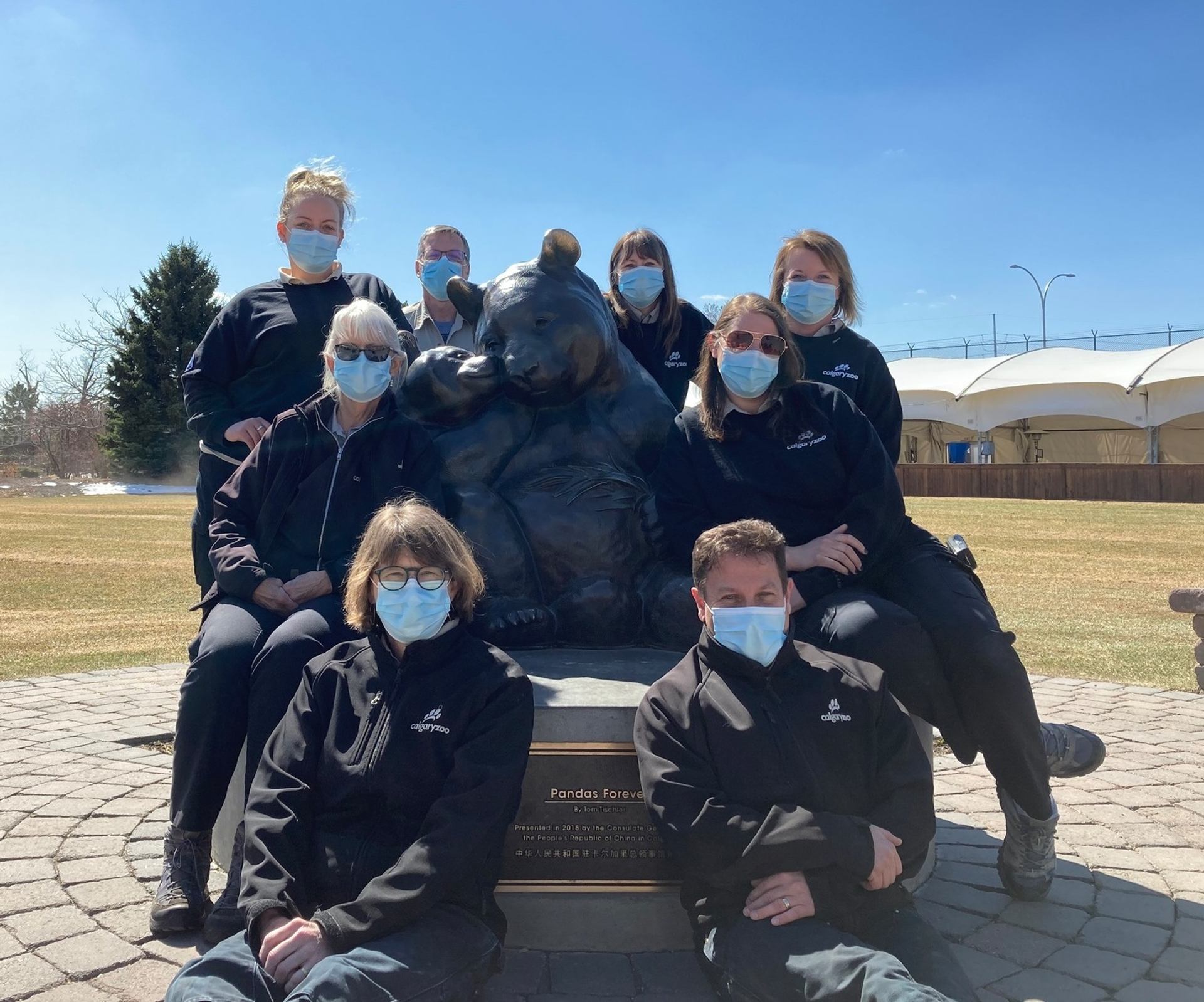 Group of masked people posing around large panda statue in outdoor park setting.
