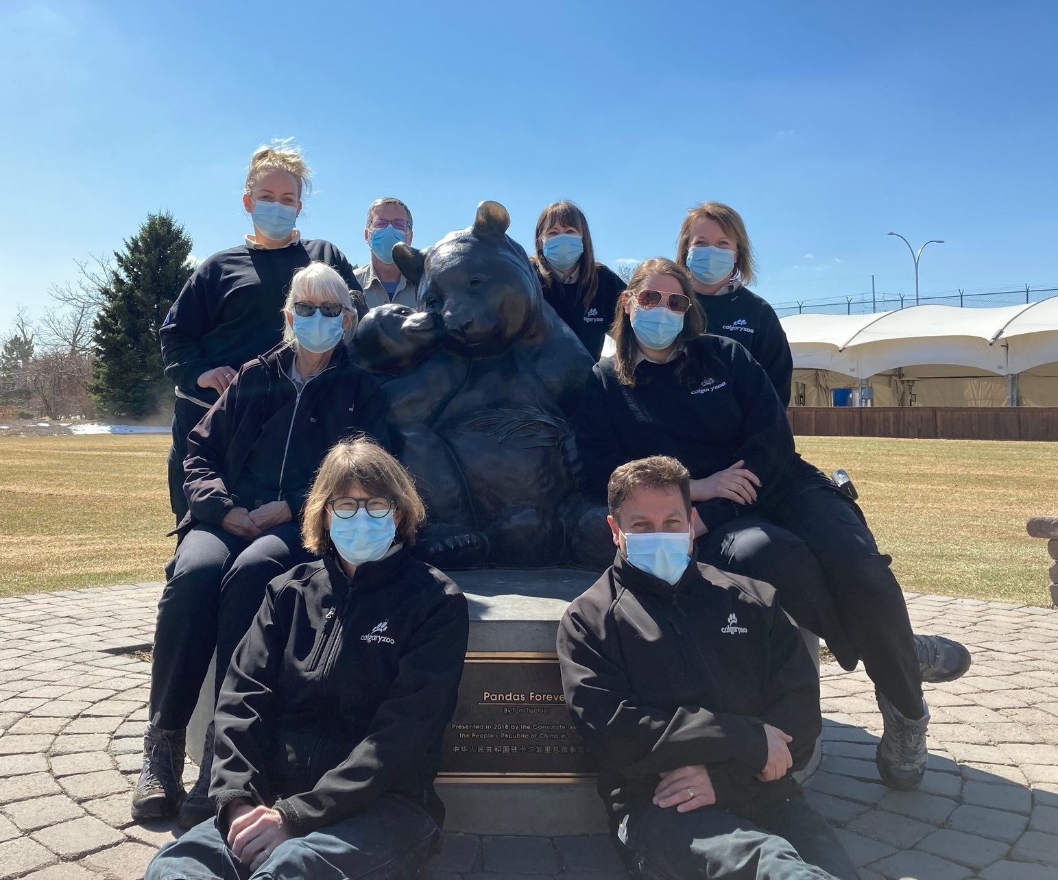 Group of masked people posing around large panda statue in outdoor park setting.