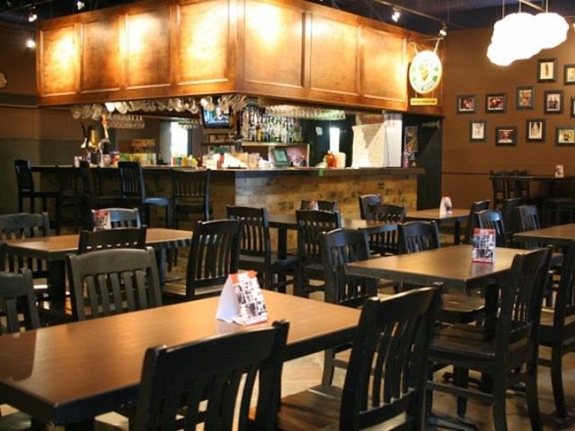 Empty restaurant dining area with wooden tables, black chairs and a bar in background.