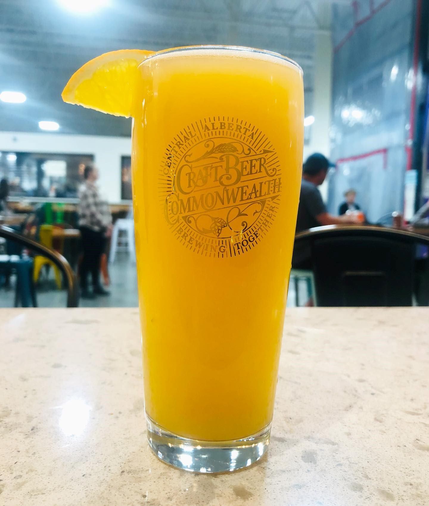 Tall glass of bright orange craft beer with an orange slice garnish on a table.