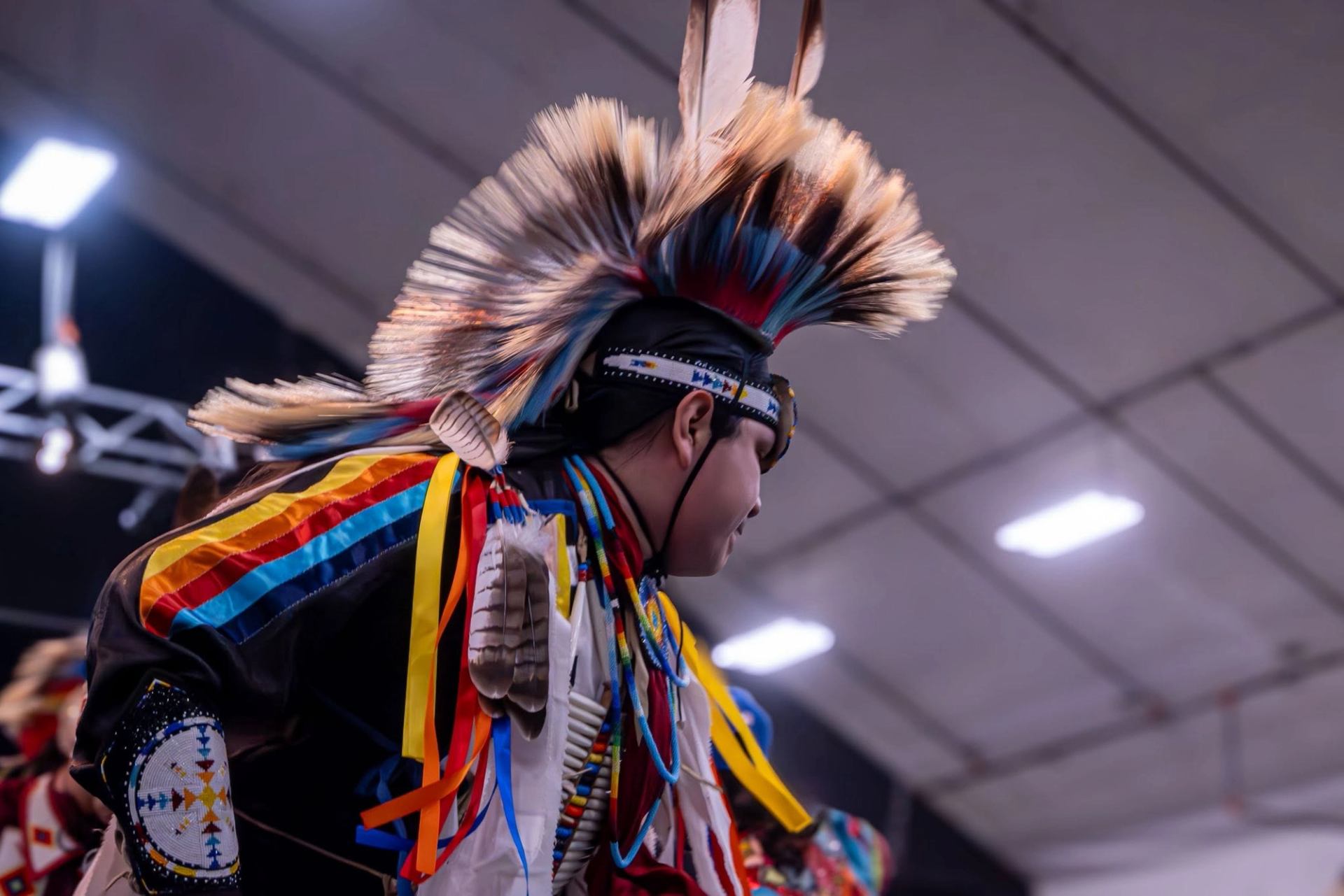 Powwow dancer in feathered headdress and colourful regalia performs indoors under bright lights.