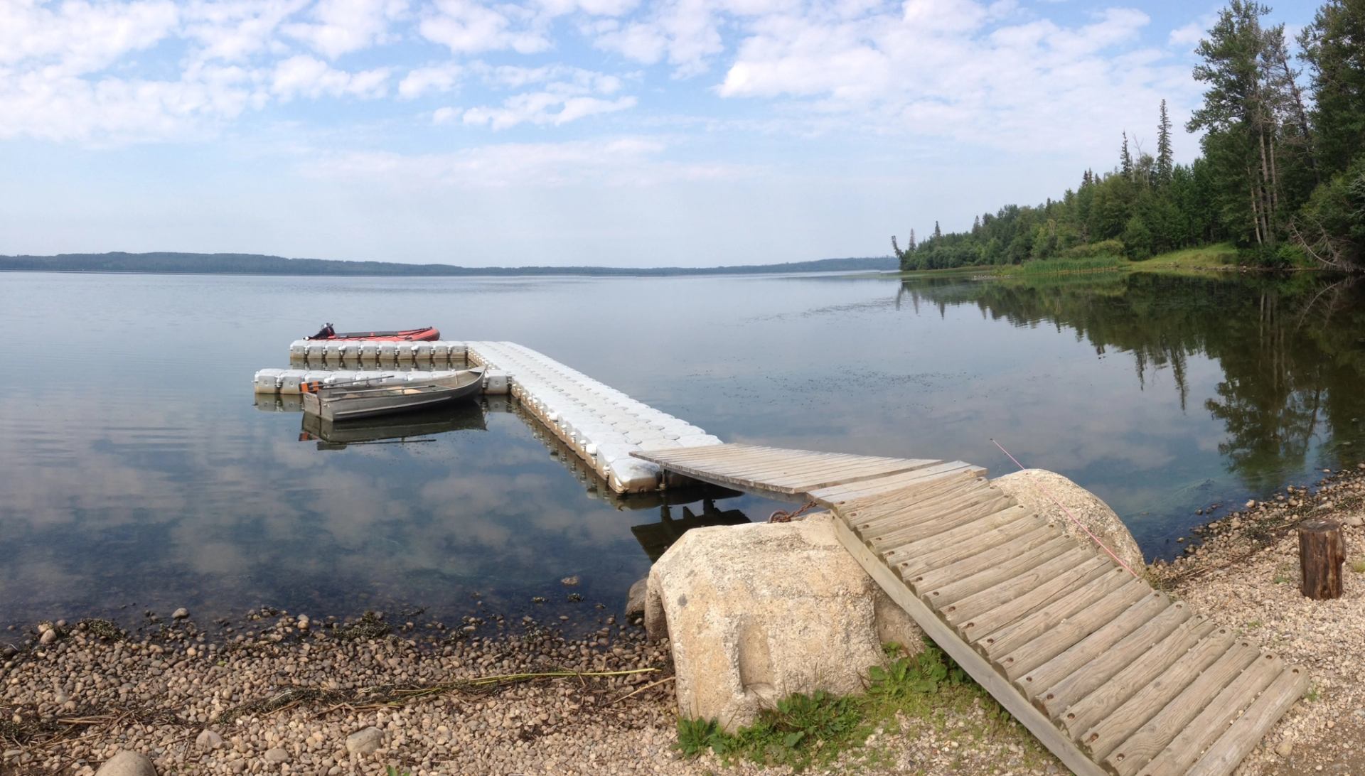 Small dock with boats on a calm lake beside a forested shoreline under a cloudy sky.
