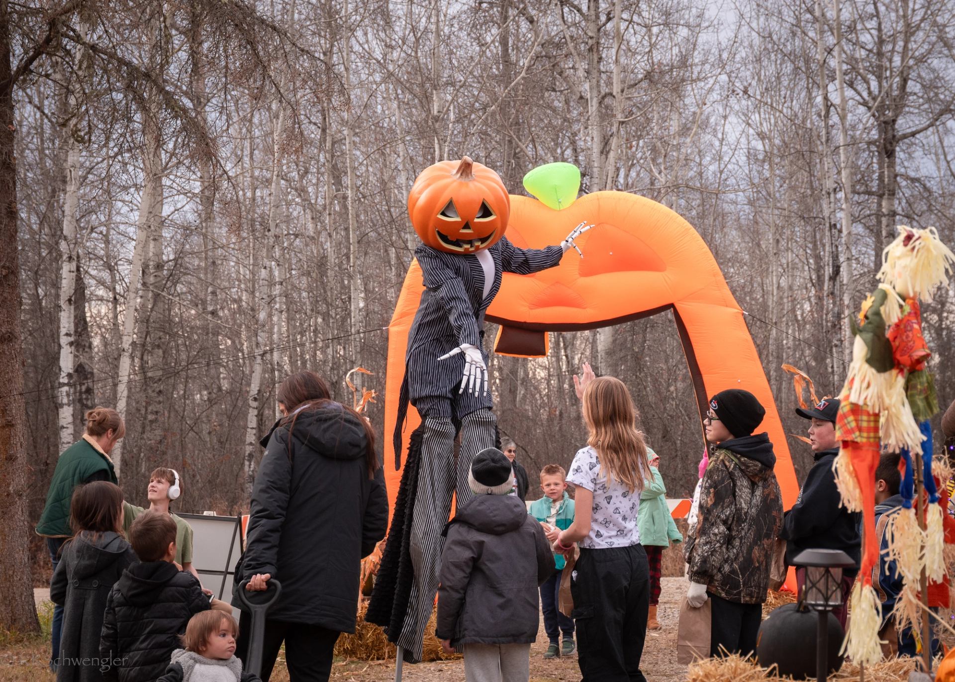 Children gather around a tall pumpkin-headed performer near an inflatable pumpkin arch.