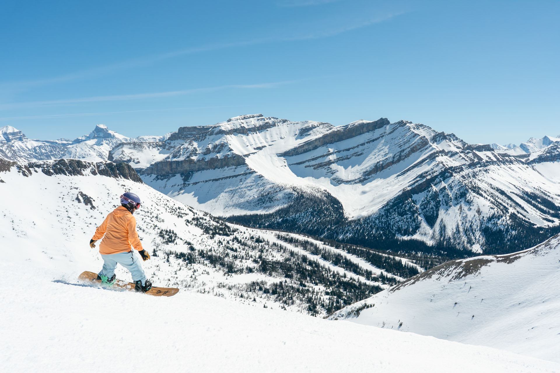 Snowboarder in orange jacket rides down snowy slope with rugged mountain range behind.
