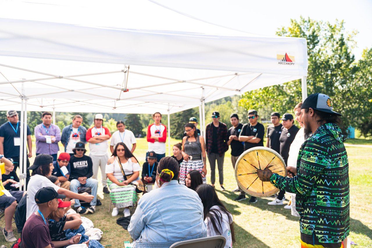 Group of people under canopy tent listening to person holding hand drum outdoors.